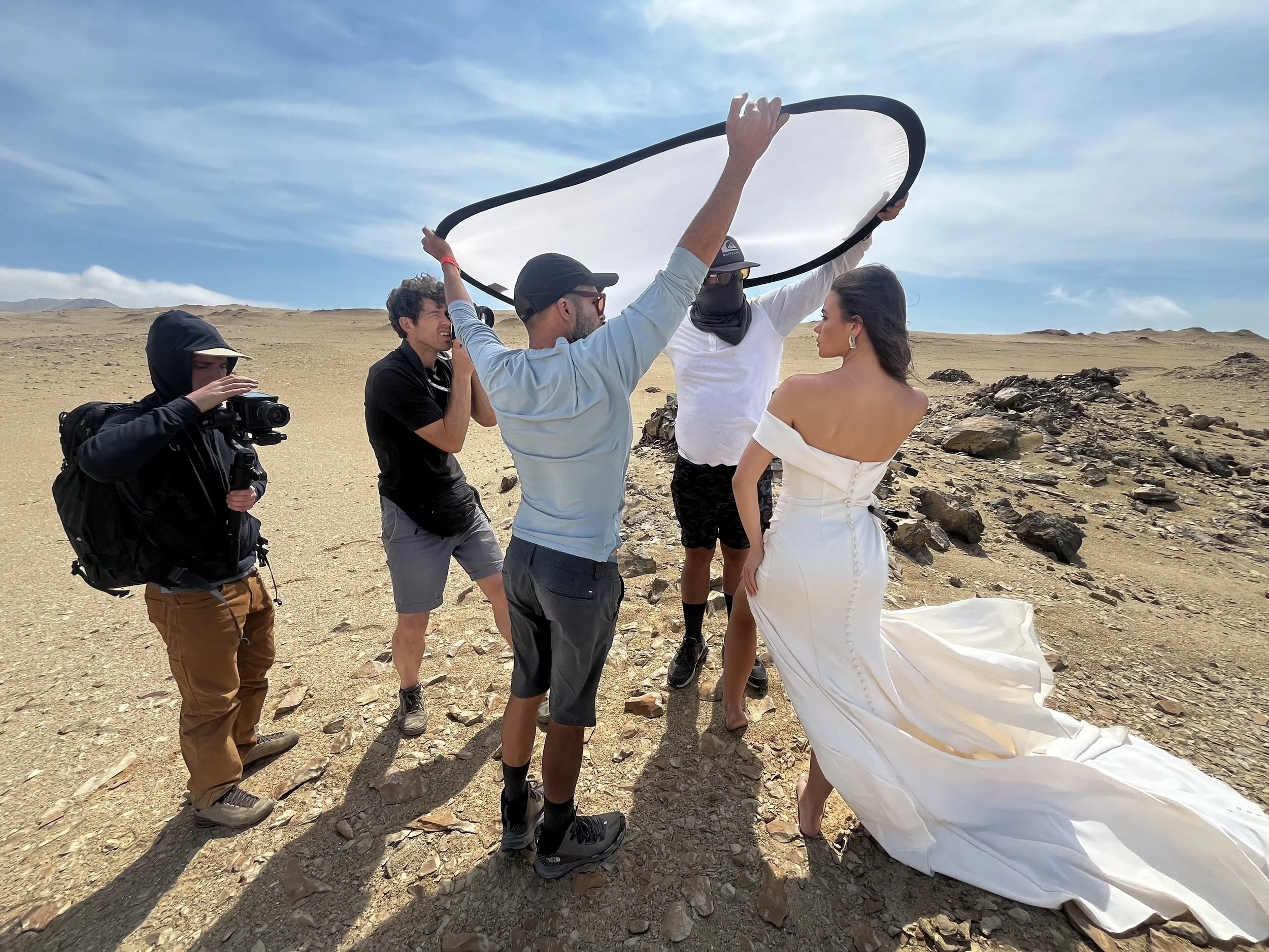 A group of people on a desert landscape during a photoshoot, with a woman in a white dress, a man holding a large reflector, a photographer, and other crew members.