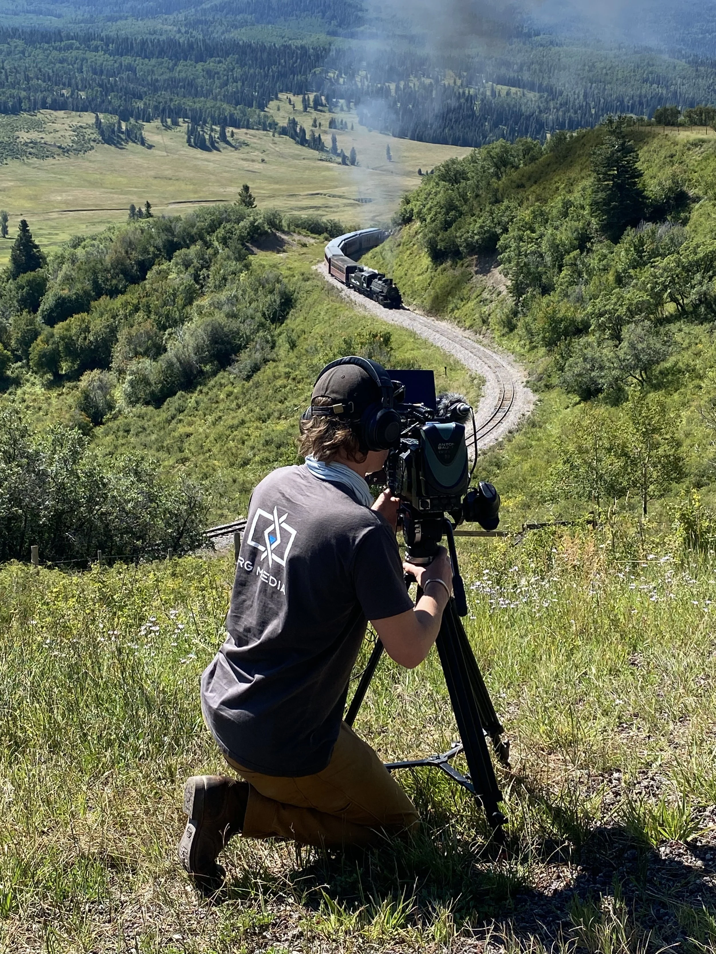 A photographer kneeling on one knee in a grassy field using a camera on a tripod to film a steam locomotive train traveling through a lush green valley surrounded by trees and hills.