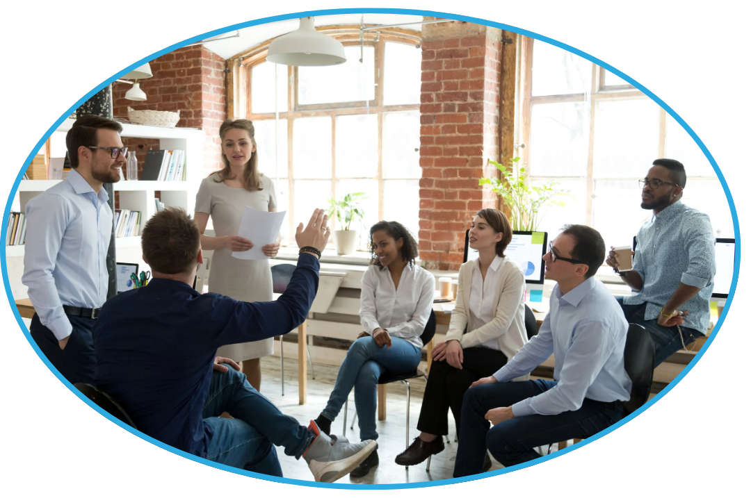 A group of professionals in a modern office setting participating in a team meeting, with some sitting and others standing, engaging in discussion.