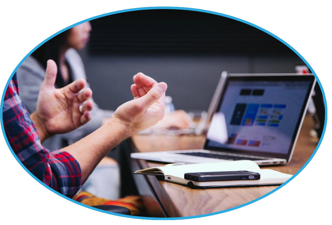 People having a discussion during a meeting, with a laptop, a smartphone, and a notebook on the table.