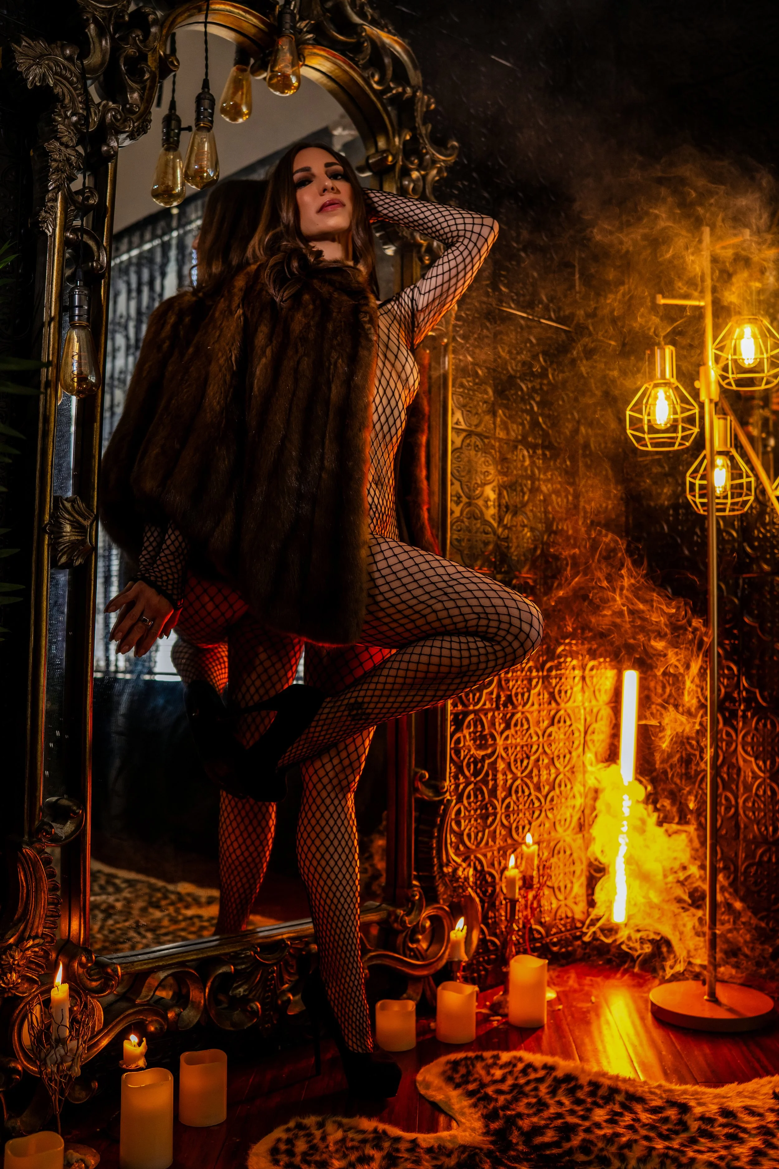 A woman with long dark hair wearing fishnet stockings, a fur coat, and high heels stands in front of a large ornate mirror. The room is decorated with candles, hanging Edison bulbs, and a decorative wall with warm lighting. Flames and smoke are visible on the right side of the image, creating a dramatic and fiery atmosphere.