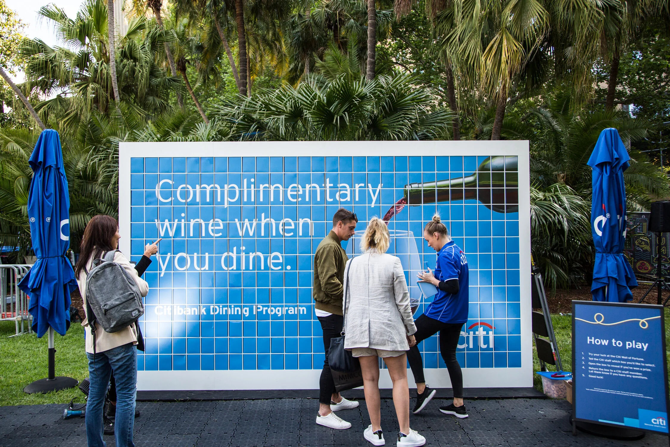 People standing in front of a large blue advertising board at an outdoor event, surrounded by palm trees and blue umbrellas.