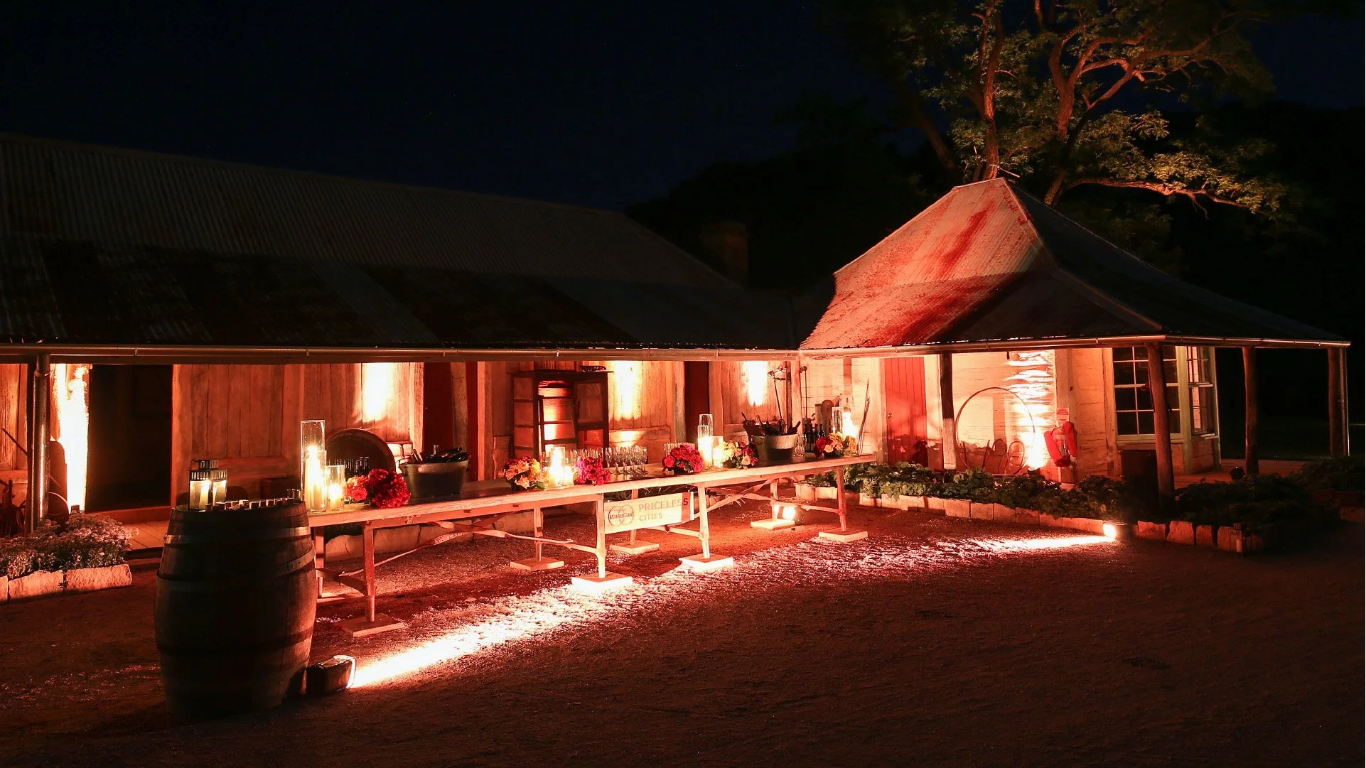 A rustic wooden building decorated with warm red lighting at night, with a long table adorned with candles and flowers in the foreground, and a large tree illuminated in the background.