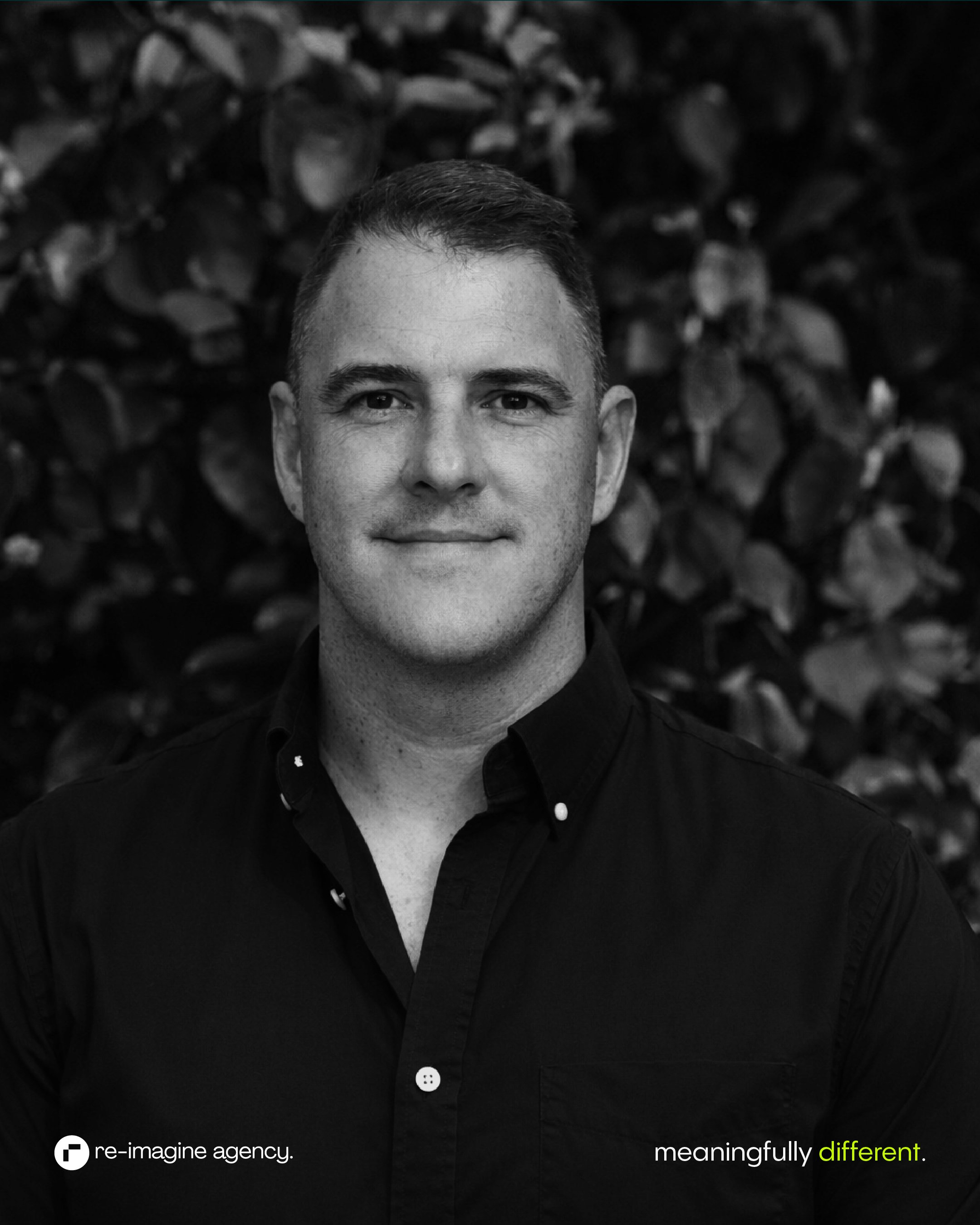 Black and white portrait of a man with short dark hair, wearing a dark collared shirt. He is standing outdoors in front of leafy bushes. The photo is focused on his face, which is centered in the frame.