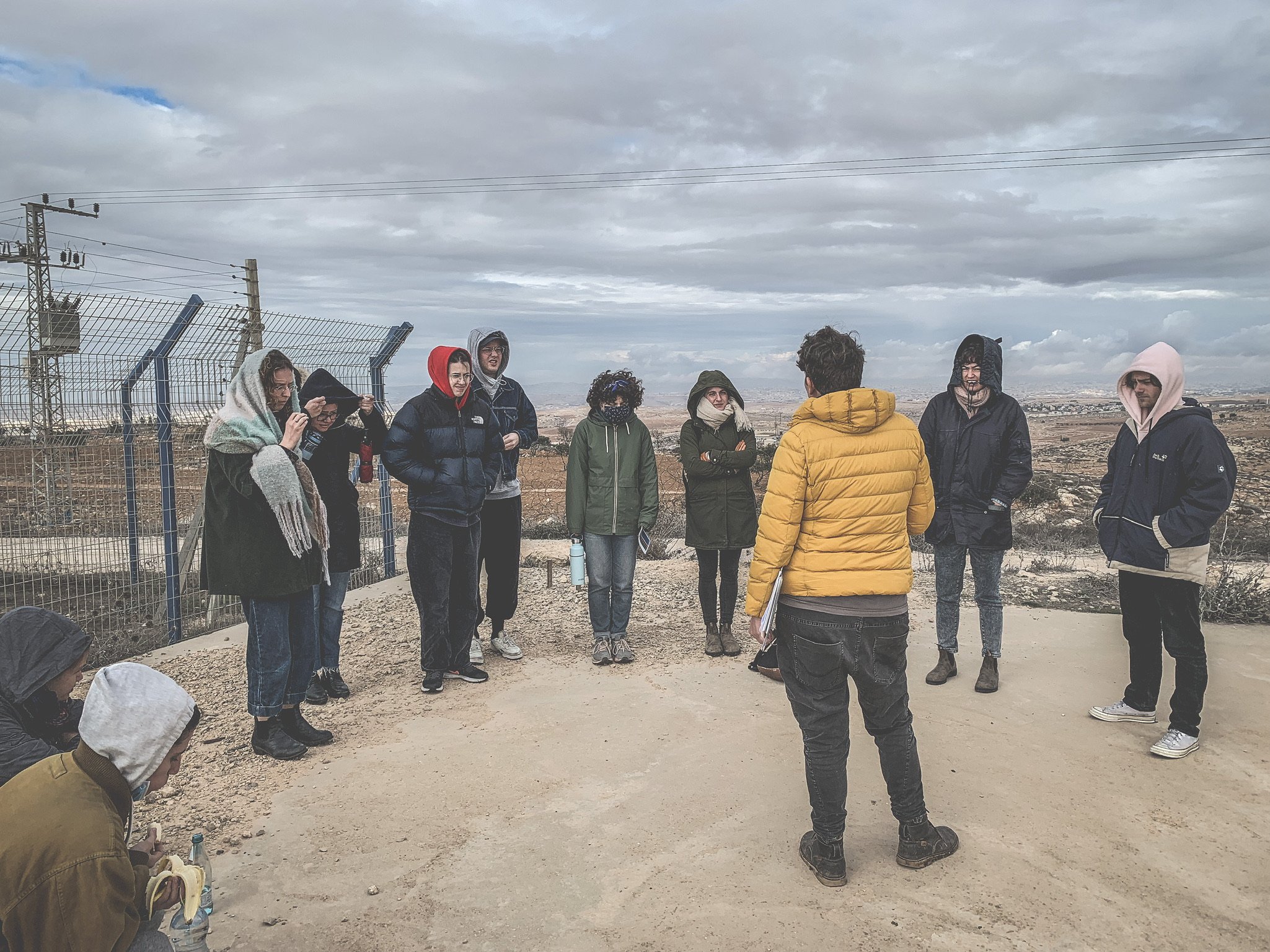  A group of people stand and listen to aperson in a yellow parka speaking in front of cloudy skies in Israel / Palestine. 