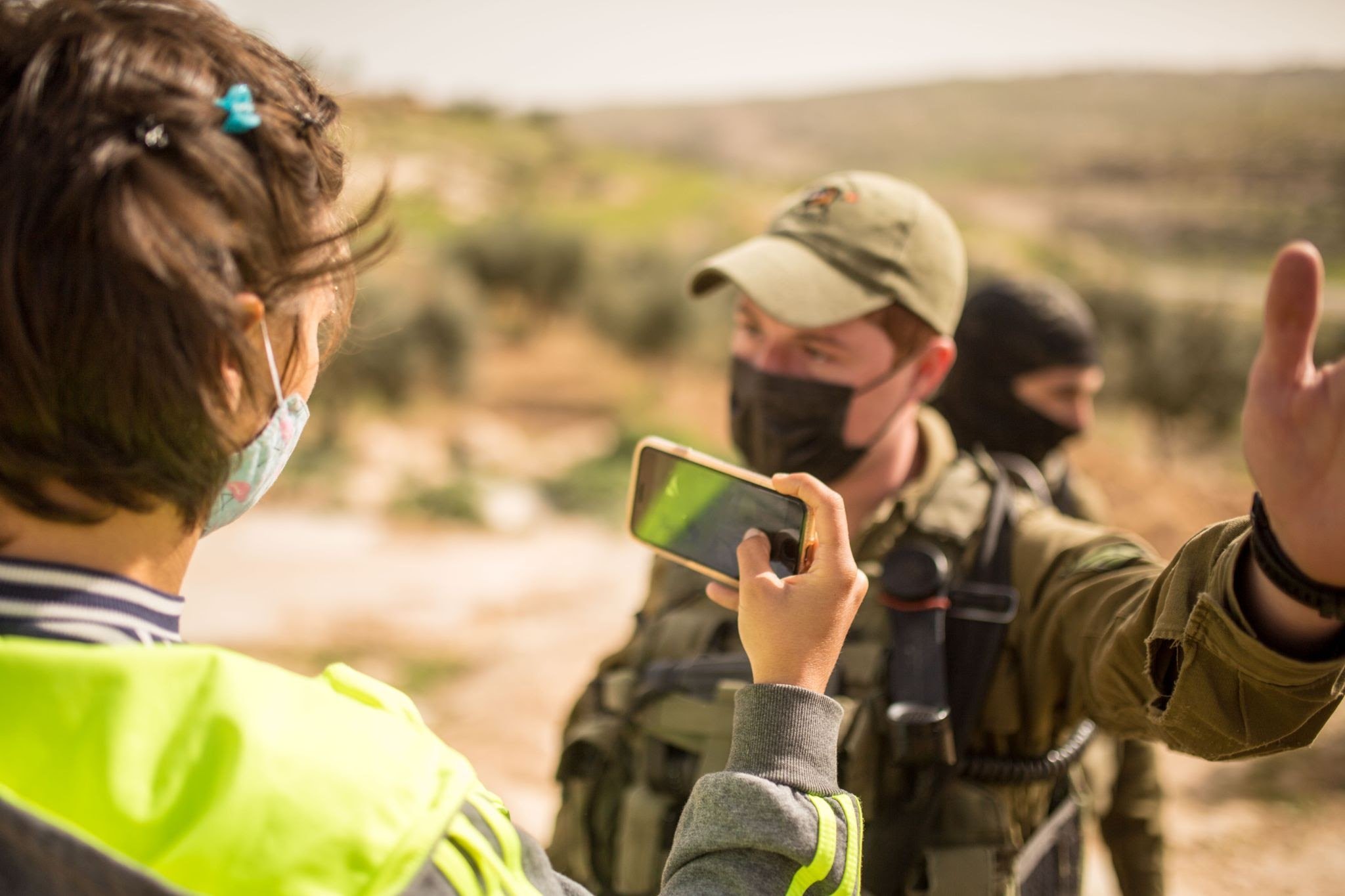  A person wearing a yellow vest holds a cell phone and documents the behaviors of a masked member of the Israeli defense force in front of them.  