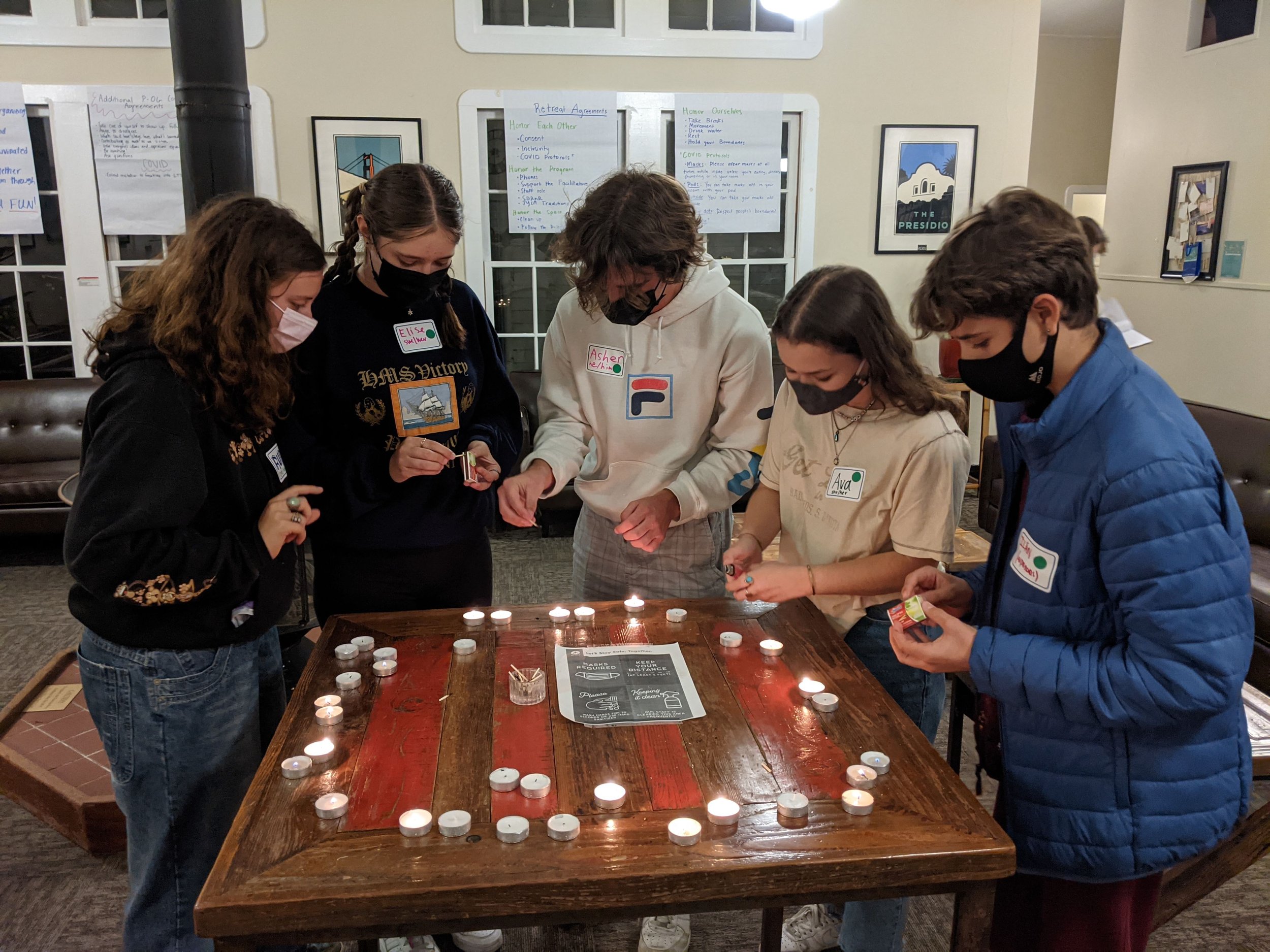  A group of young people wearing masks and lighting Shabbat candles around a table 