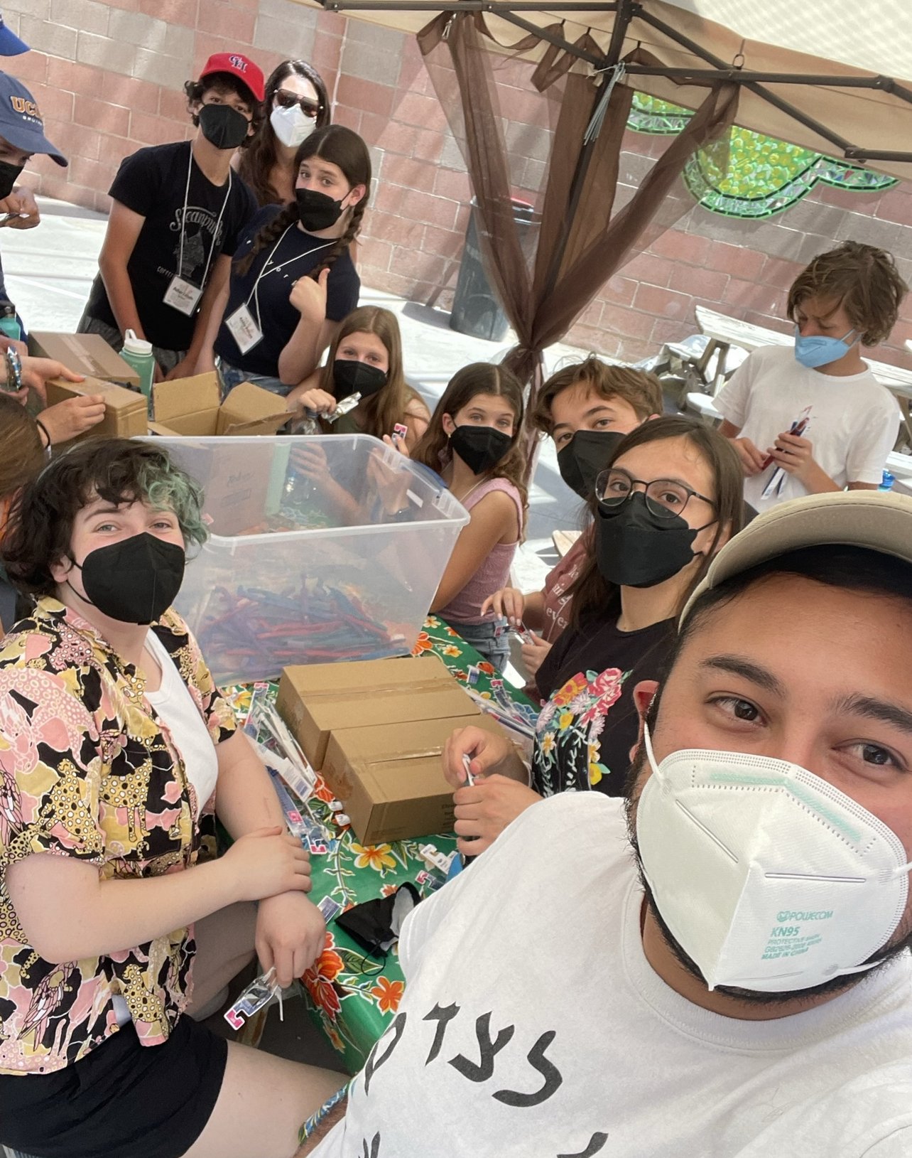  A selfie taken by a man wearing a hat of a group of people wearing masks packing items into a storage container and make as if they’re smiling.  
