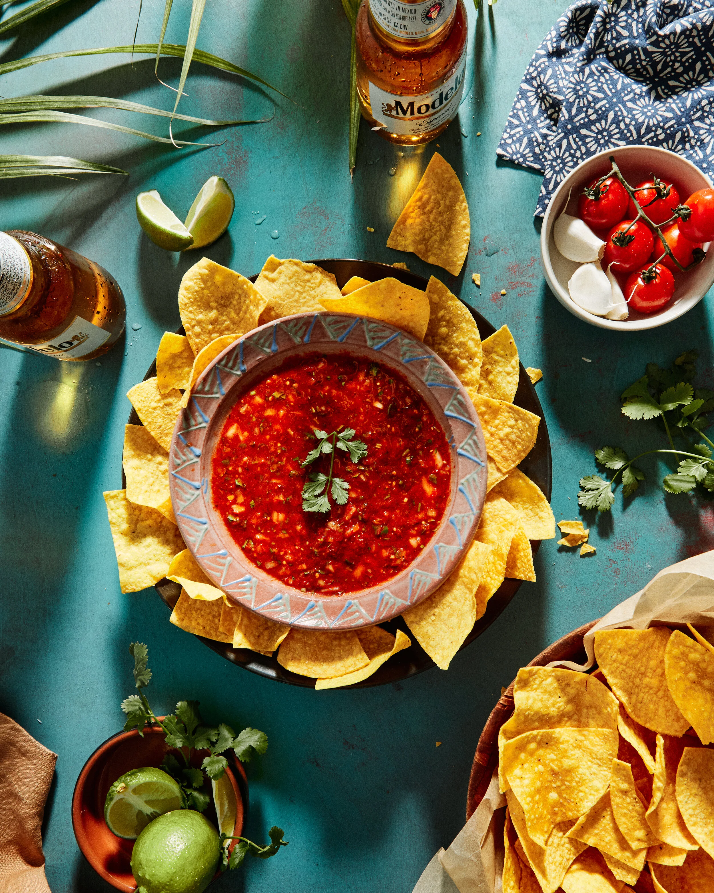 Golden corn tortilla chips on a platter with a bowl of chili de arbol salsa. Dining table in kitchen scene in Mexico with limes, Modelo cerveza, and fresh produce.