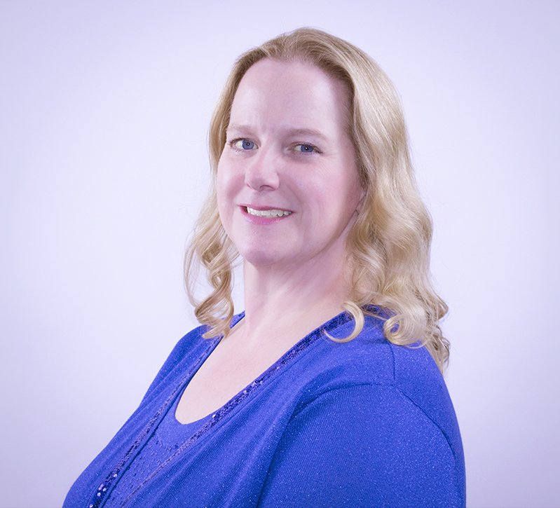 A woman with blonde hair wearing a blue top, smiling with a neutral background.