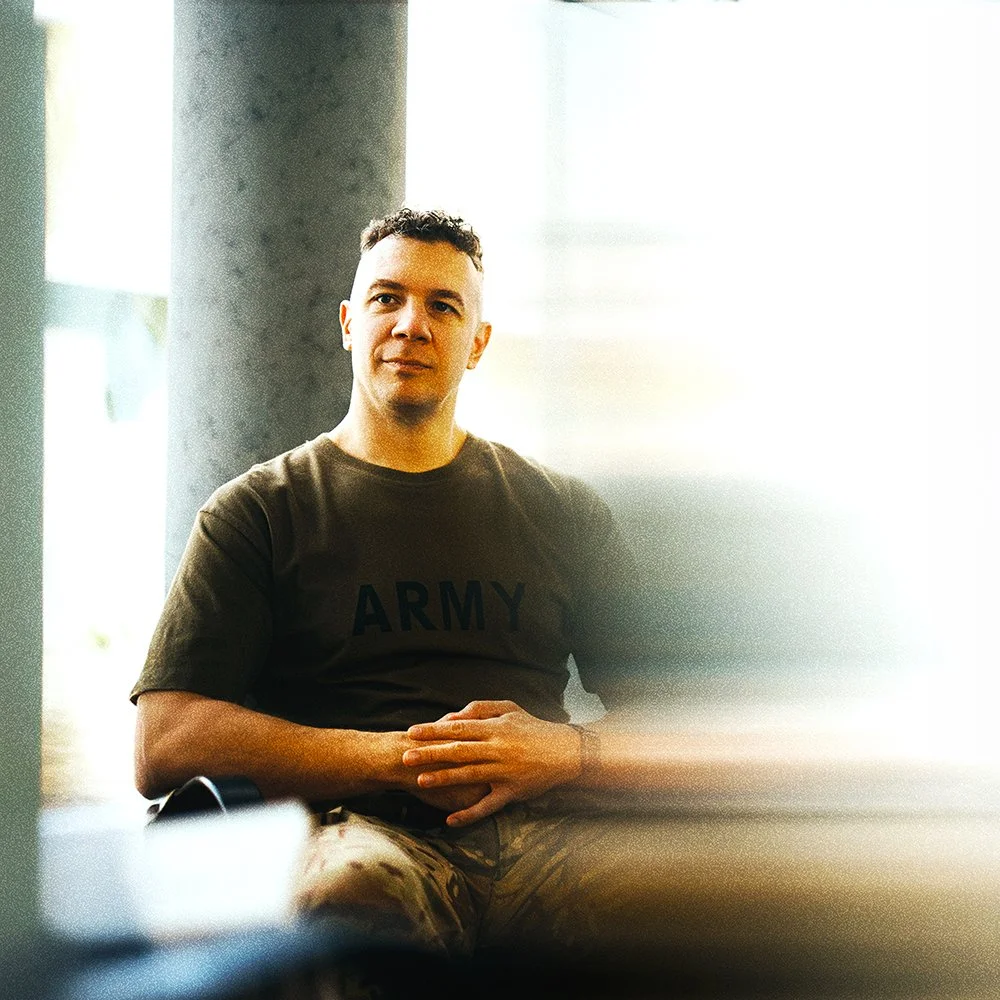 A man in military uniform sitting indoors near a large window, wearing a black shirt with
