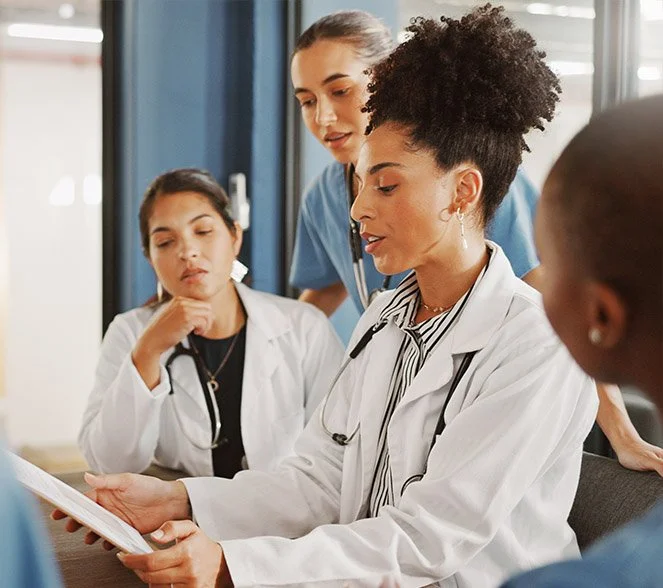Four diverse medical professionals, including a physician with curly hair and three others, gathered around a document in a hospital or clinic setting, engaged in discussion or review.