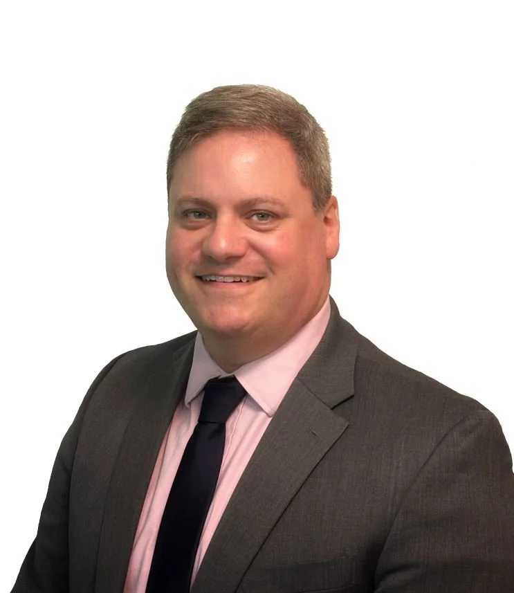 Professional headshot of a man in a dark gray suit, pink dress shirt, and black tie, smiling at the camera