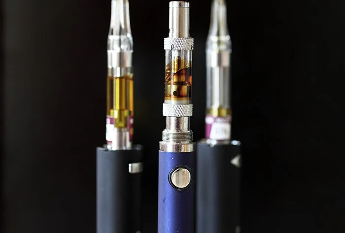 Three vape pens with cartridges filled with liquid nicotine or flavored vapor, standing upright against a dark background.