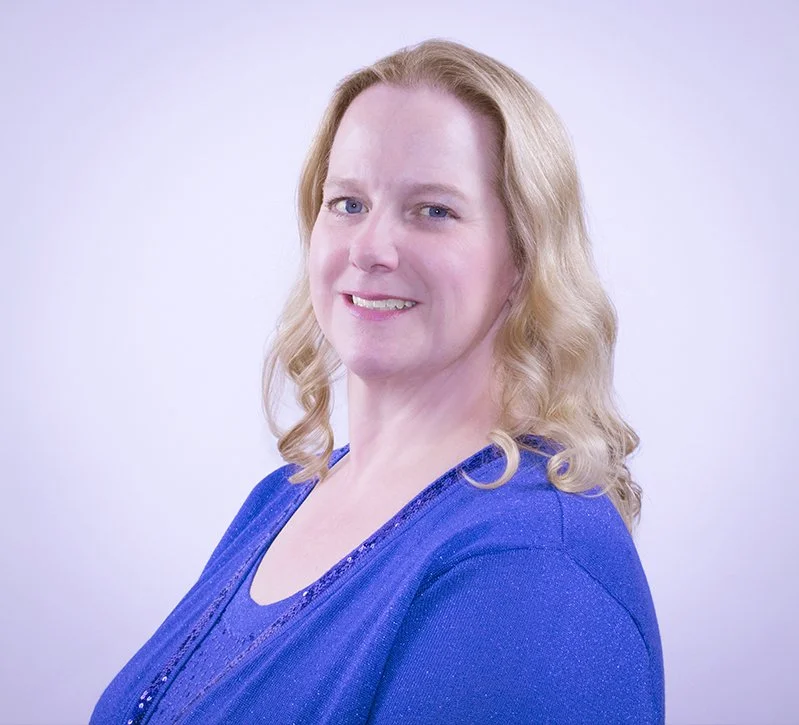 A woman with blonde curly hair smiling at the camera, wearing a blue top, against a light background.