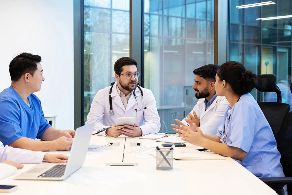 A group of medical professionals, including doctors and nurses, having a meeting in a clinical office with large windows, discussing patient care or medical topics.