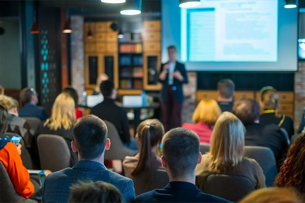 A group of people attending a presentation or seminar in a conference room with a speaker standing at the front near a large screen.