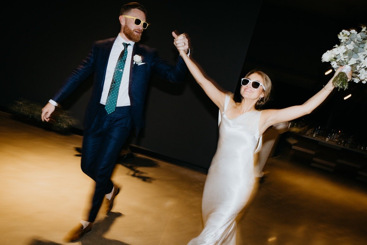 A wedding couple dancing, the man in a navy suit with a white shirt, teal tie, and sunglasses, holding hands with the woman in a white dress and sunglasses, holding a bouquet of flowers, in a dimly lit indoor setting.