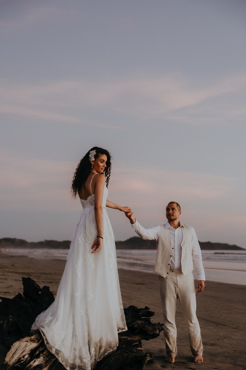 A wedding in Santa Teresa, the bride in a white wedding dress standing on a piece of driftwood on the beach, holding hands with a man in white pants and a vest, with the ocean and a partly cloudy sky in the background during sunset.
