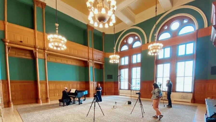 Musicians practicing in a large, elegant room with high arched windows and ornate chandeliers, setup with microphones and cameras.