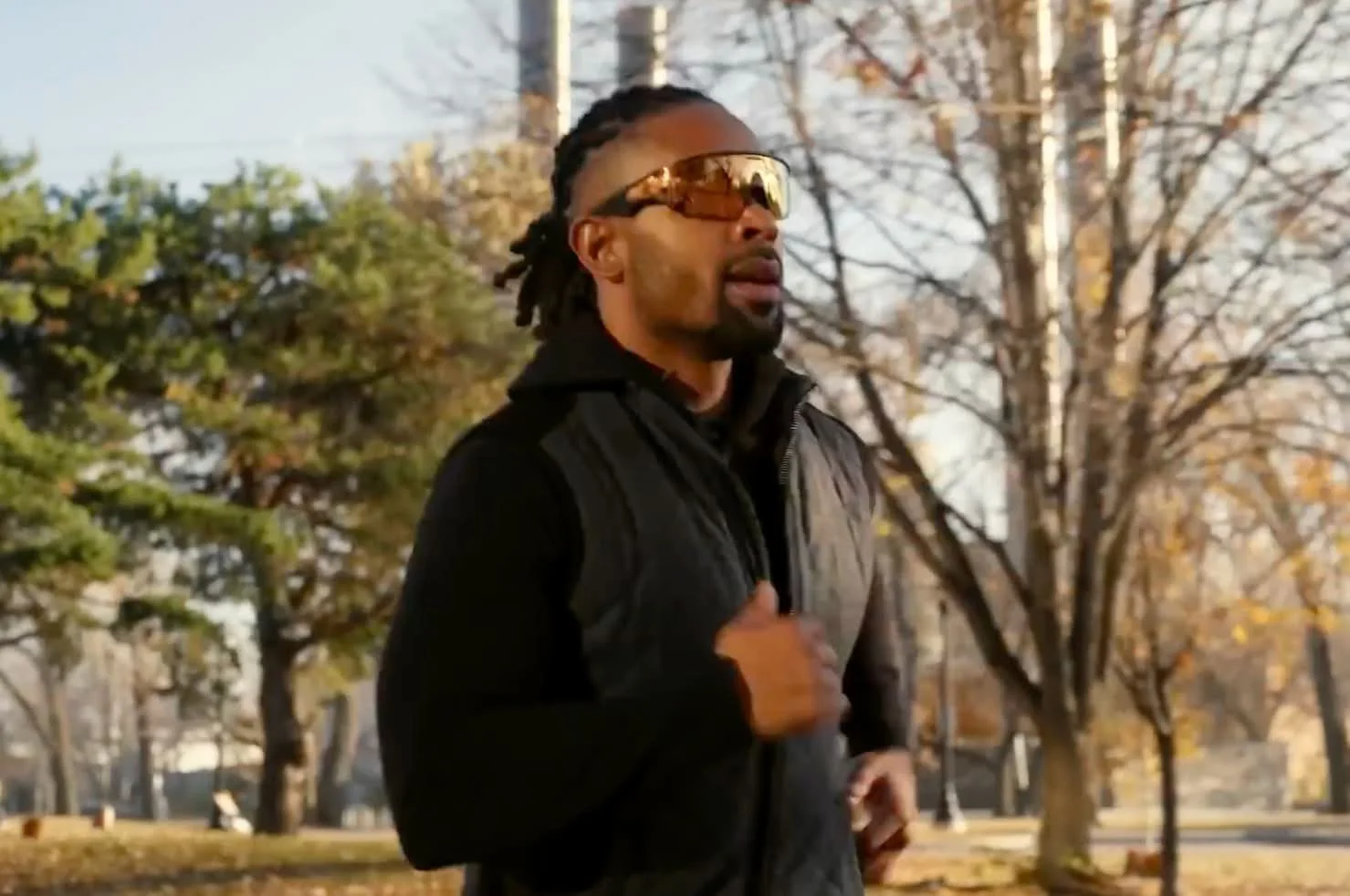 Man jogging outdoors in a park with trees and autumn foliage, wearing sunglasses and a black jacket.