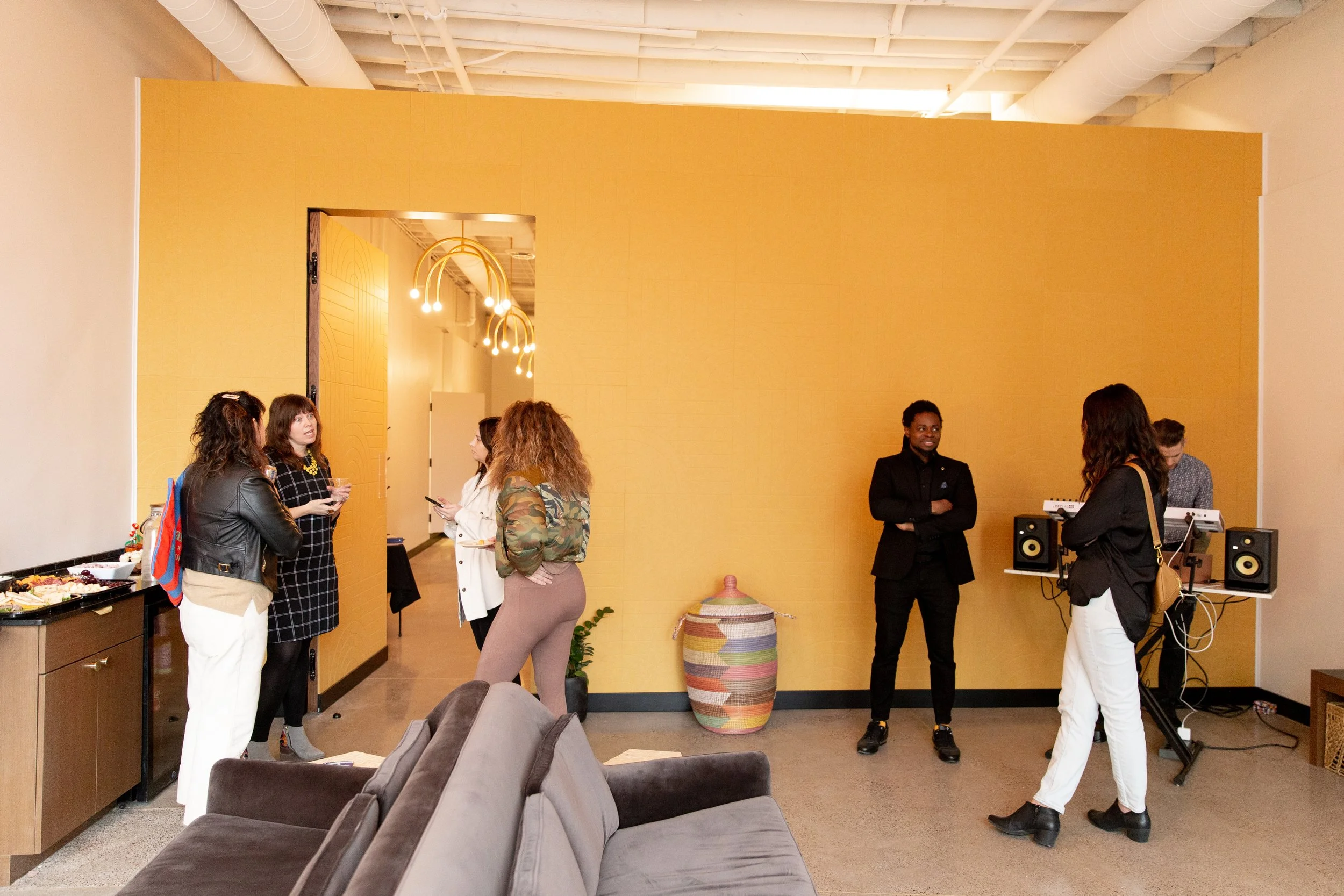 People socializing in a brightly lit room with yellow walls, a doorway, and musical speakers in the corner. Some guests are holding drinks and engaging in conversation, with a table of food visible on the left side.