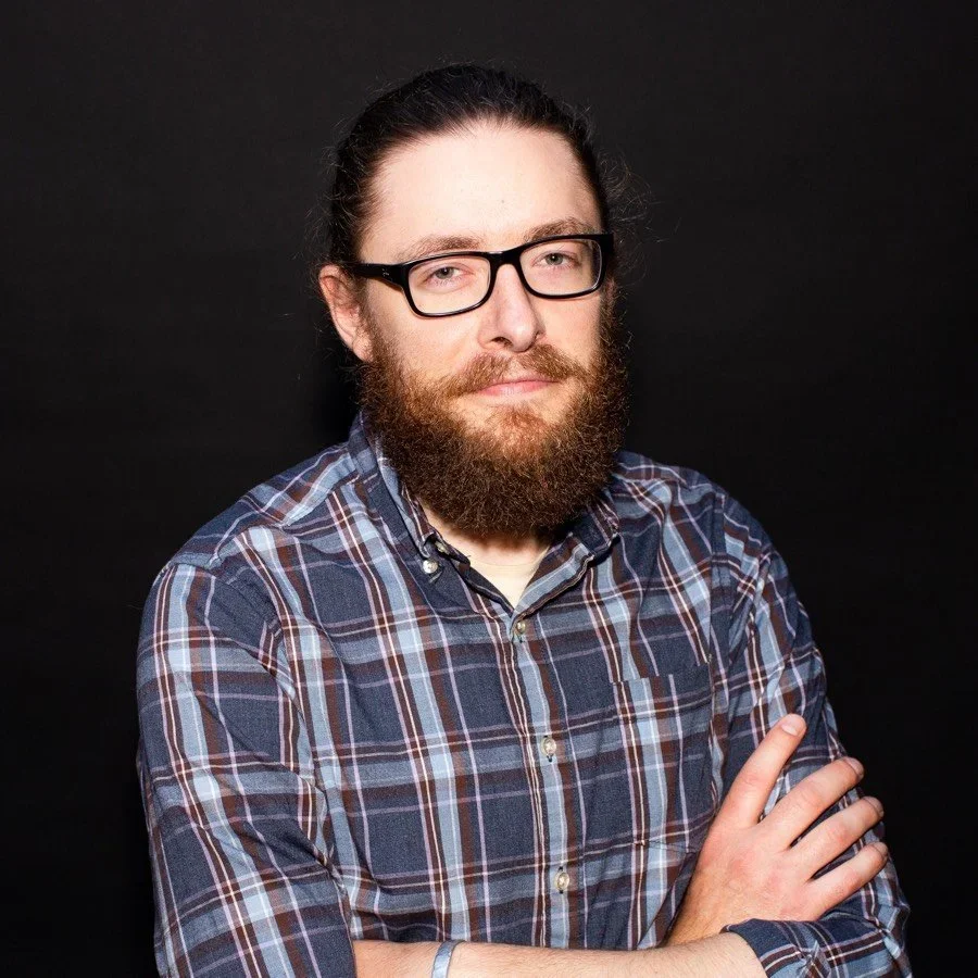 Man with long hair, glasses, and a beard wearing a plaid shirt, standing in front of a black background.
