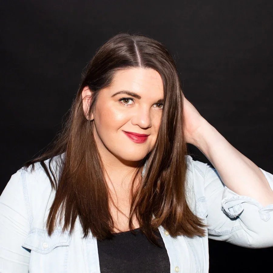 A woman with long brown hair, wearing a white shirt over a black top, posing against a black background.