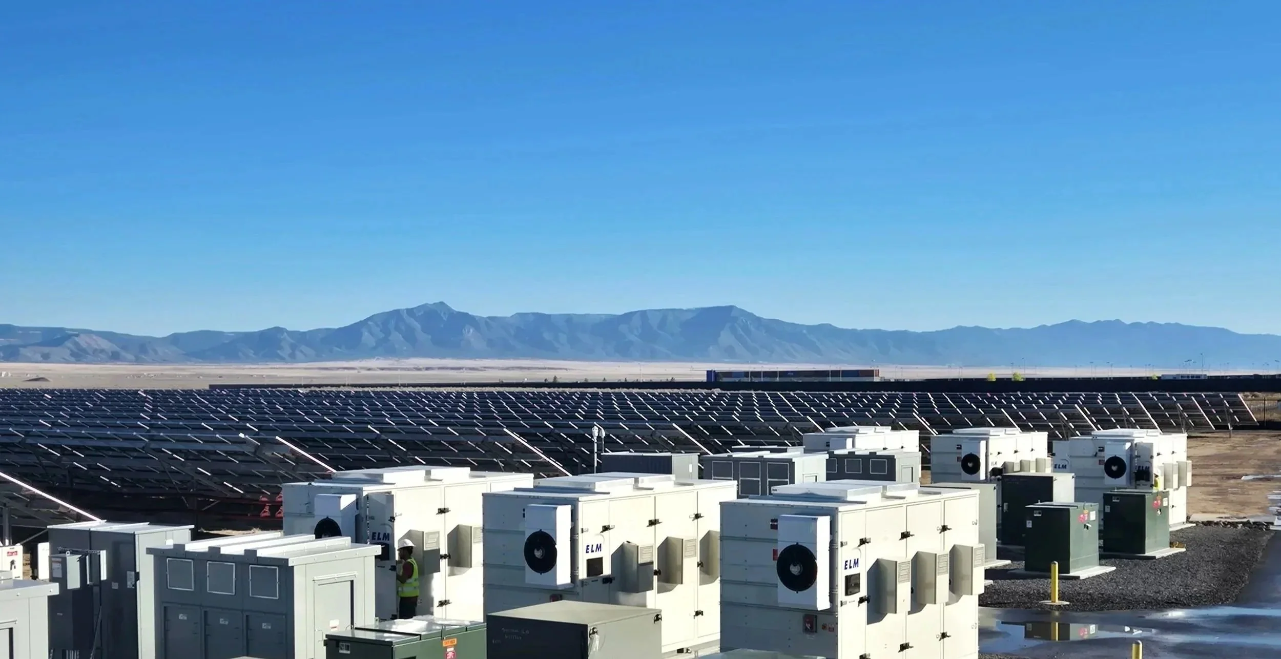 A utility scale solar array with numerous solar panels, and an energy storage system, set against a backdrop of mountains and clear blue sky in Albuquerque New Mexico.