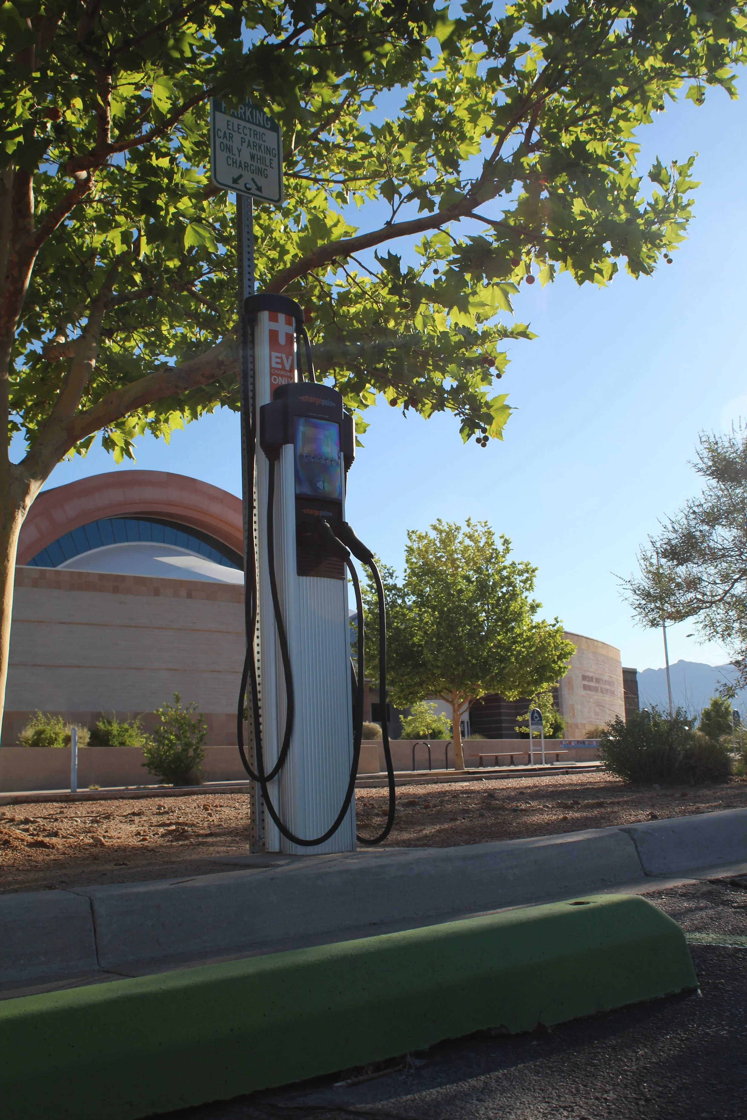 Electric vehicle charging station with a sign indicating parking only while charging, trees in the background, and a building with an arched roof under a clear blue sky.