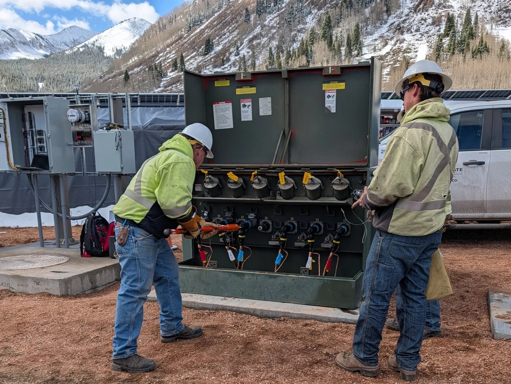Two workers in safety gear inspecting and working on electrical equipment outdoors with mountainous snow-covered landscape in the background.