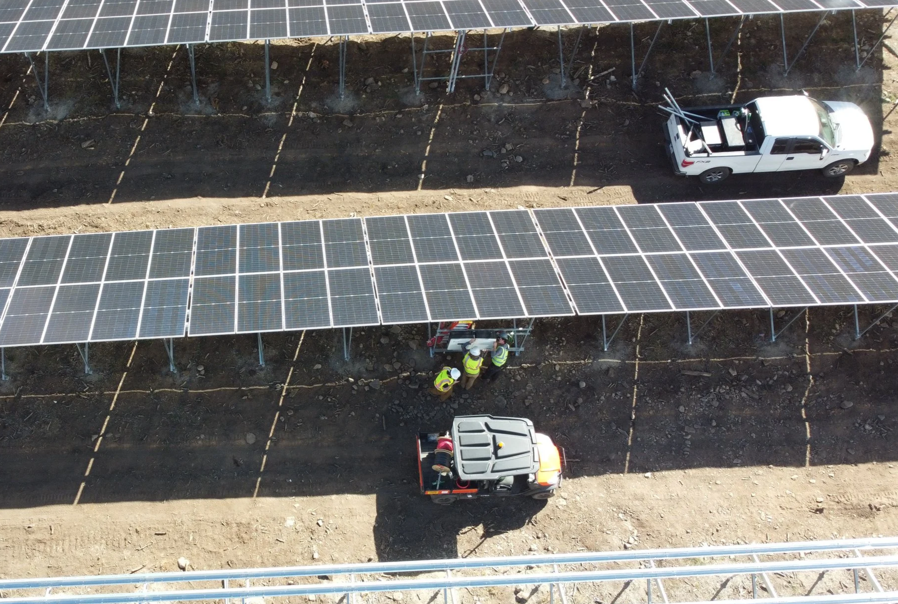 Aerial view of utility scale solar panel installation on a dirt field, with workers and trucks involved in setup.
