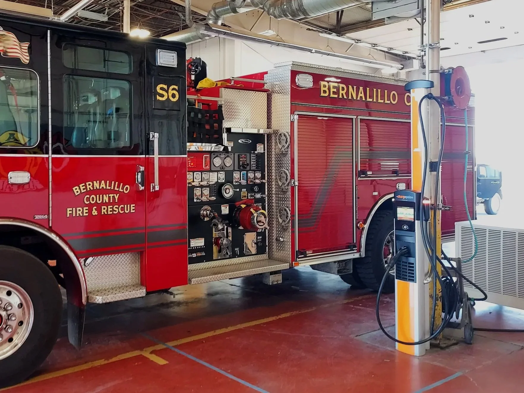 A large red fire truck labeled 'Bernalillo County Fire & Rescue' is parked inside a fire station with an electric vehicle charging station nearby.