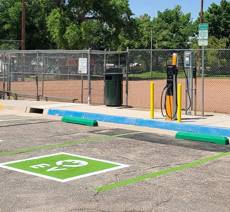 Electric vehicle charging station at a parking lot with a designated green electric vehicle parking space in the foreground, surrounded by a fence and trees.