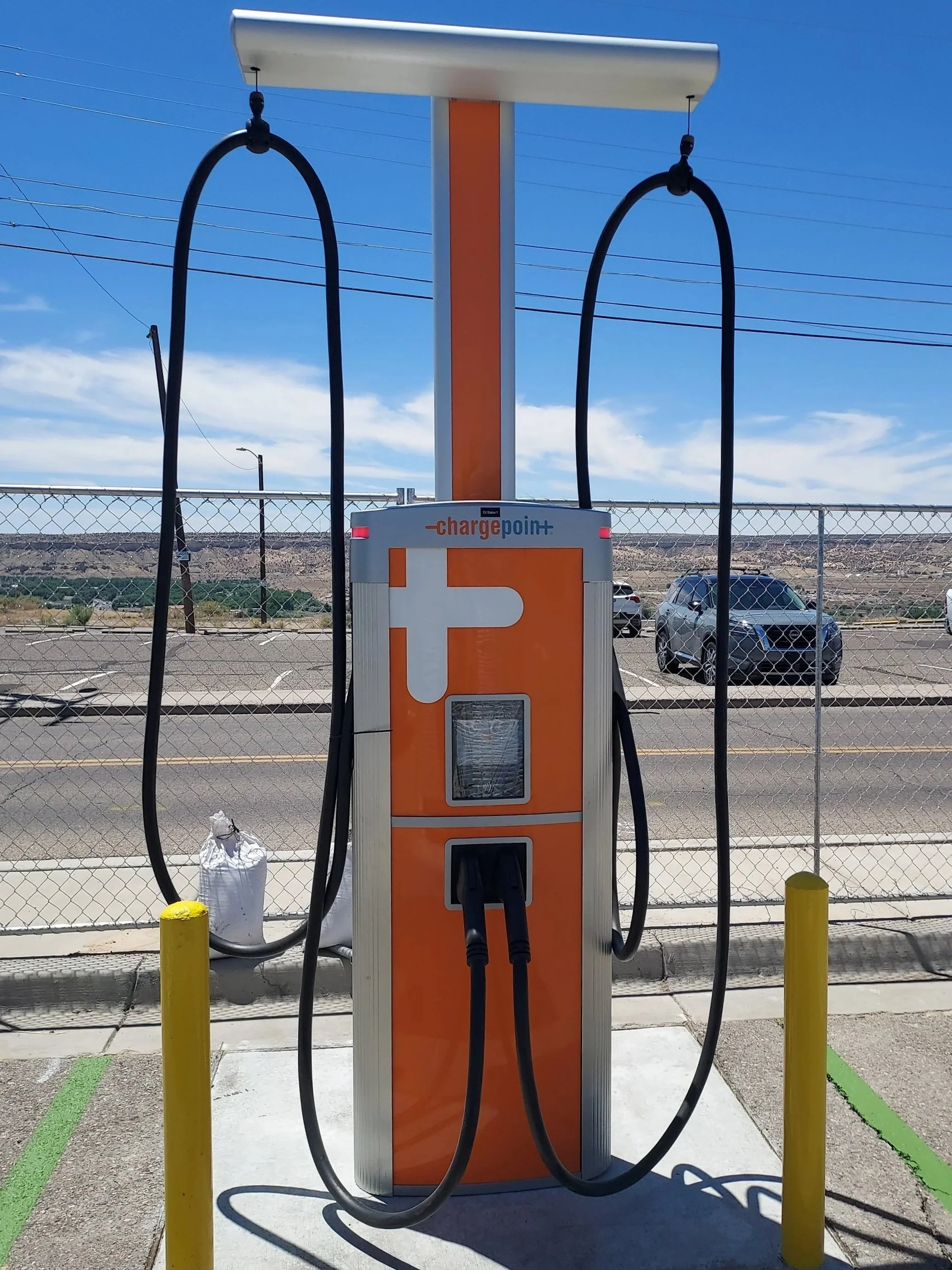 Electric vehicle charging station with two charging cables, located outdoors in a parking lot with a chain-link fence and parked cars in the background.