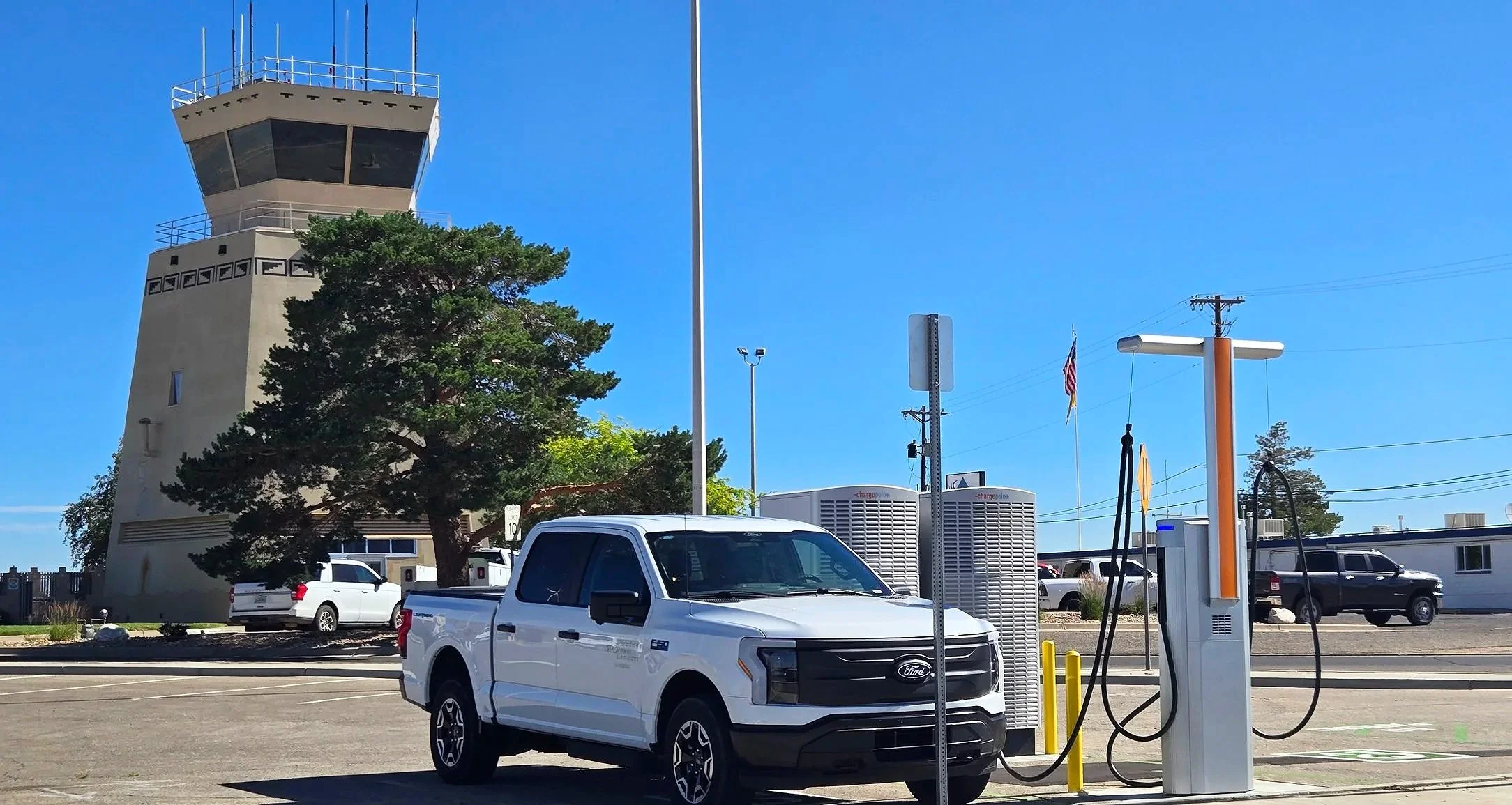 A white electric pickup truck parked at an electric vehicle charging station in a parking lot under a clear blue sky, with an air traffic control tower in the background.