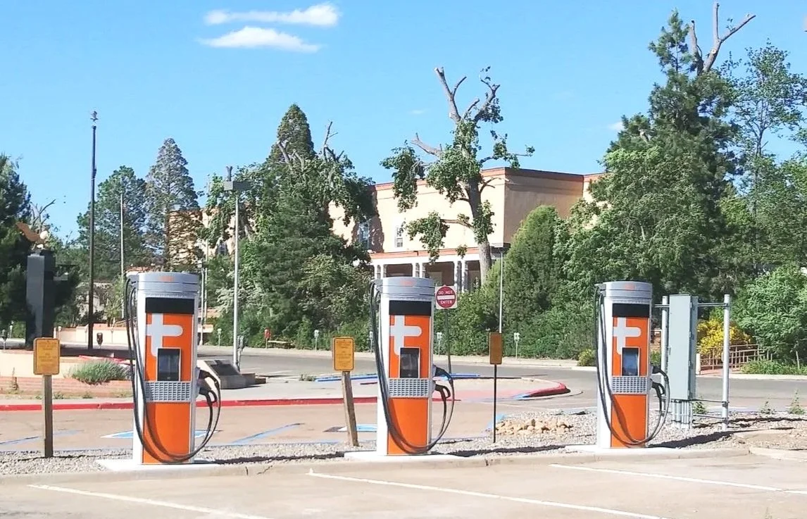 Three electric vehicle charging stations in a parking lot with trees and a building in the background.