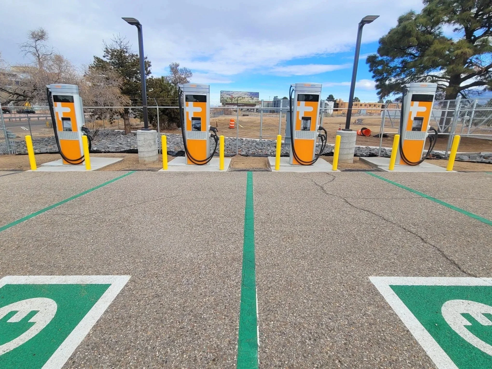 Four electric vehicle charging stations in a NMDOT parking lot. There are trees and construction equipment behind a chain-link fence.