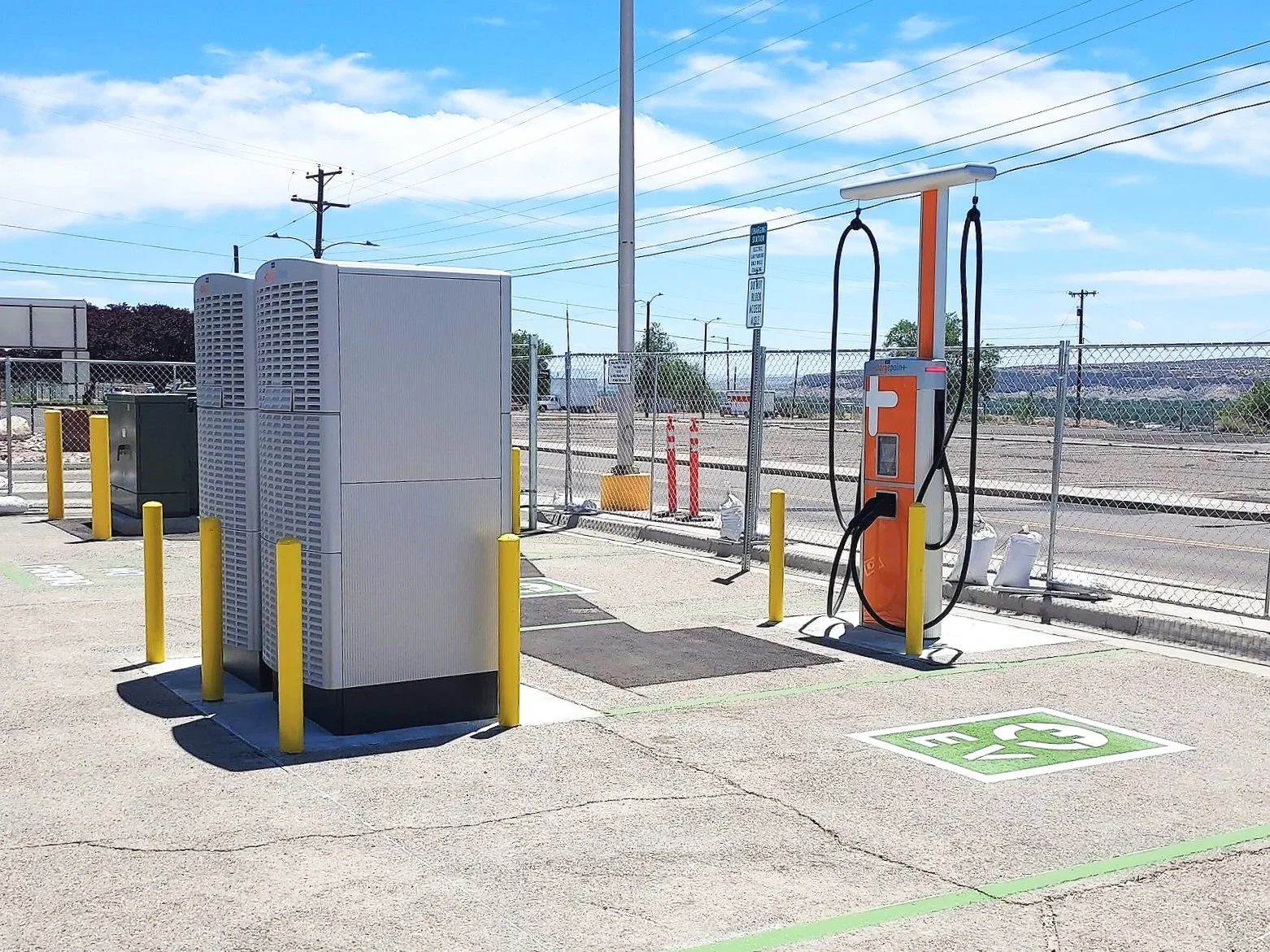 Electric vehicle charging station in a parking lot at the Four Corners Regional Airport with utility equipment and a chain-link fence in the background under a partly cloudy sky.