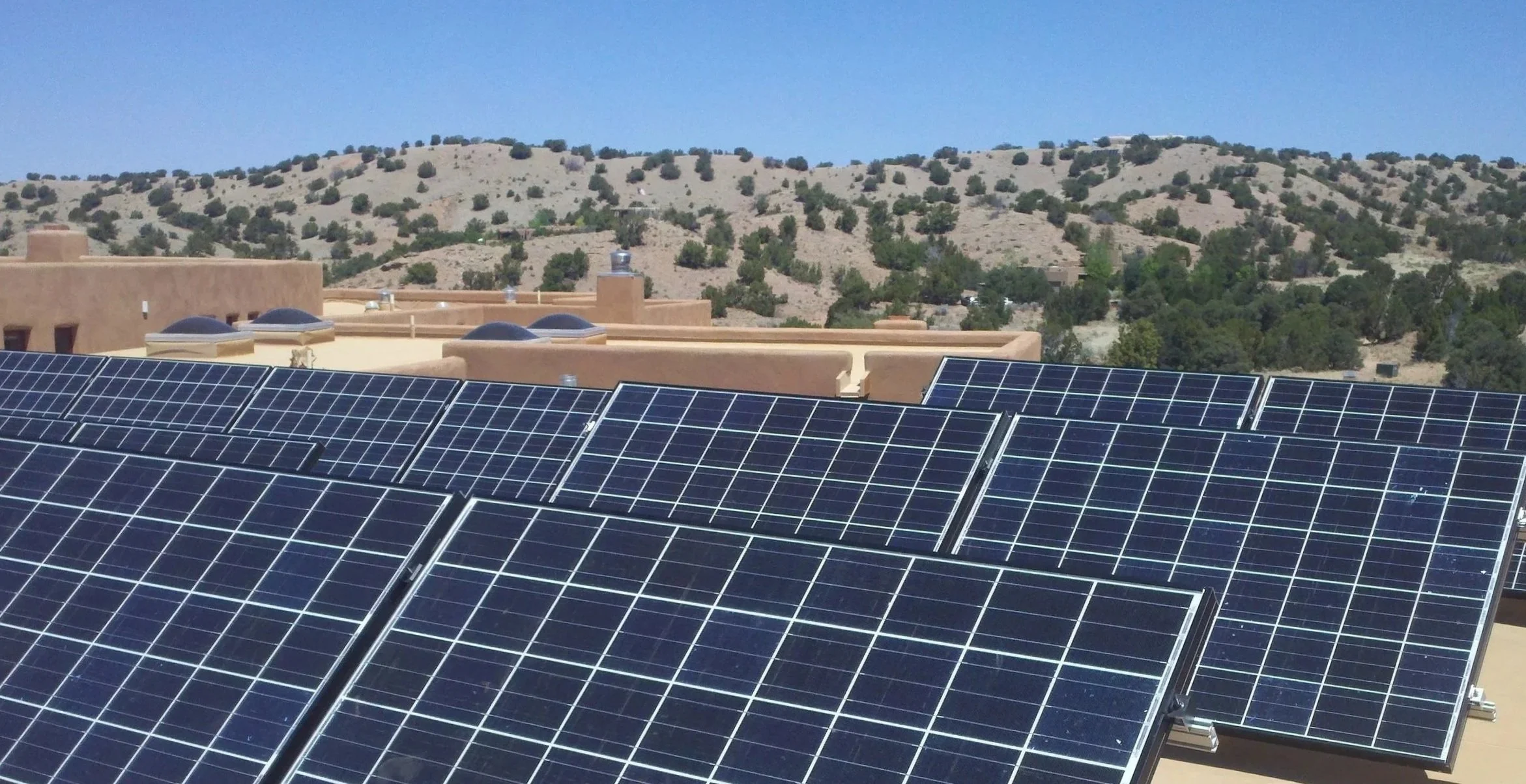 Multiple solar panels installed on a residential rooftop with a desert landscape and hills in the background.