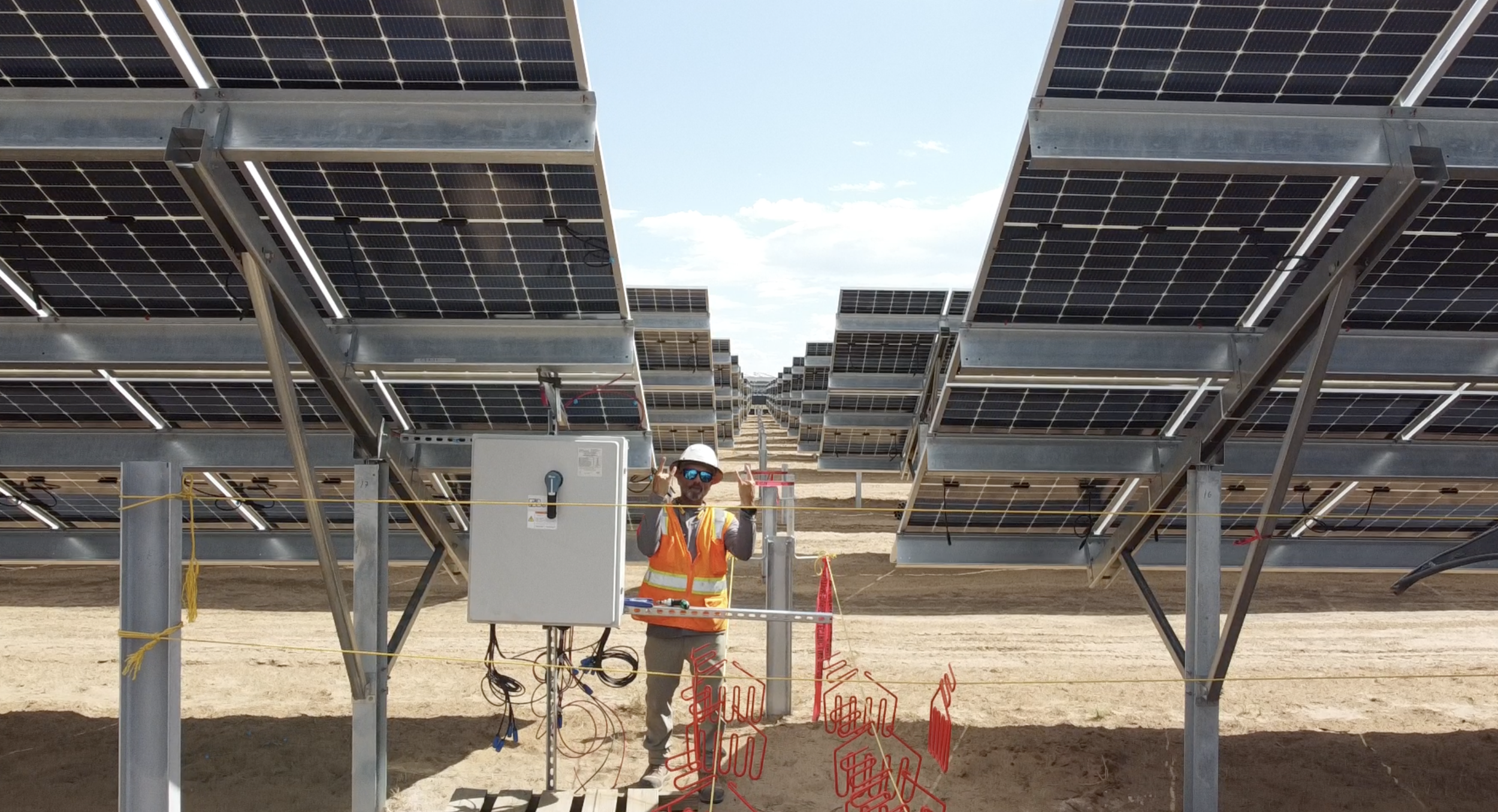 A worker wearing a safety helmet, sunglasses, and a reflective vest standing among solar panel arrays at a solar energy installation site, with a clear blue sky overhead.