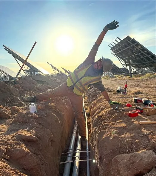 A construction worker in a safety vest and hard hat smiling and waving while working in a trench with pipes at a solar farm site during sunset.
