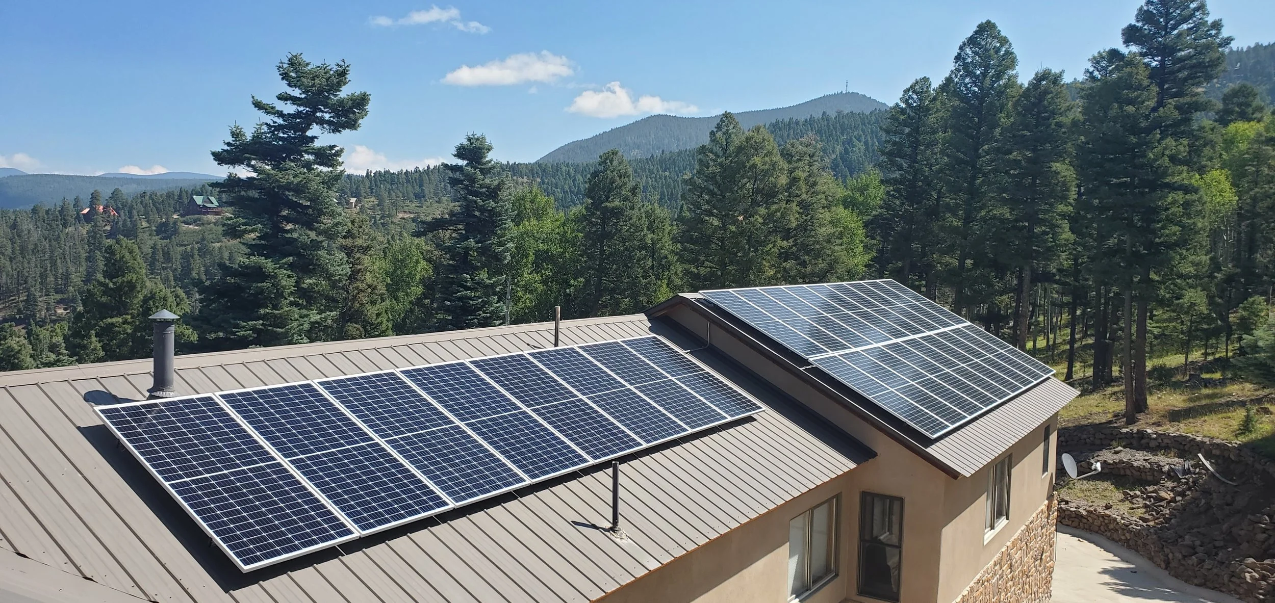 Solar panels installed on the roof of a house in a mountainous wooded area under a blue sky in Angel Fire New Mexico.