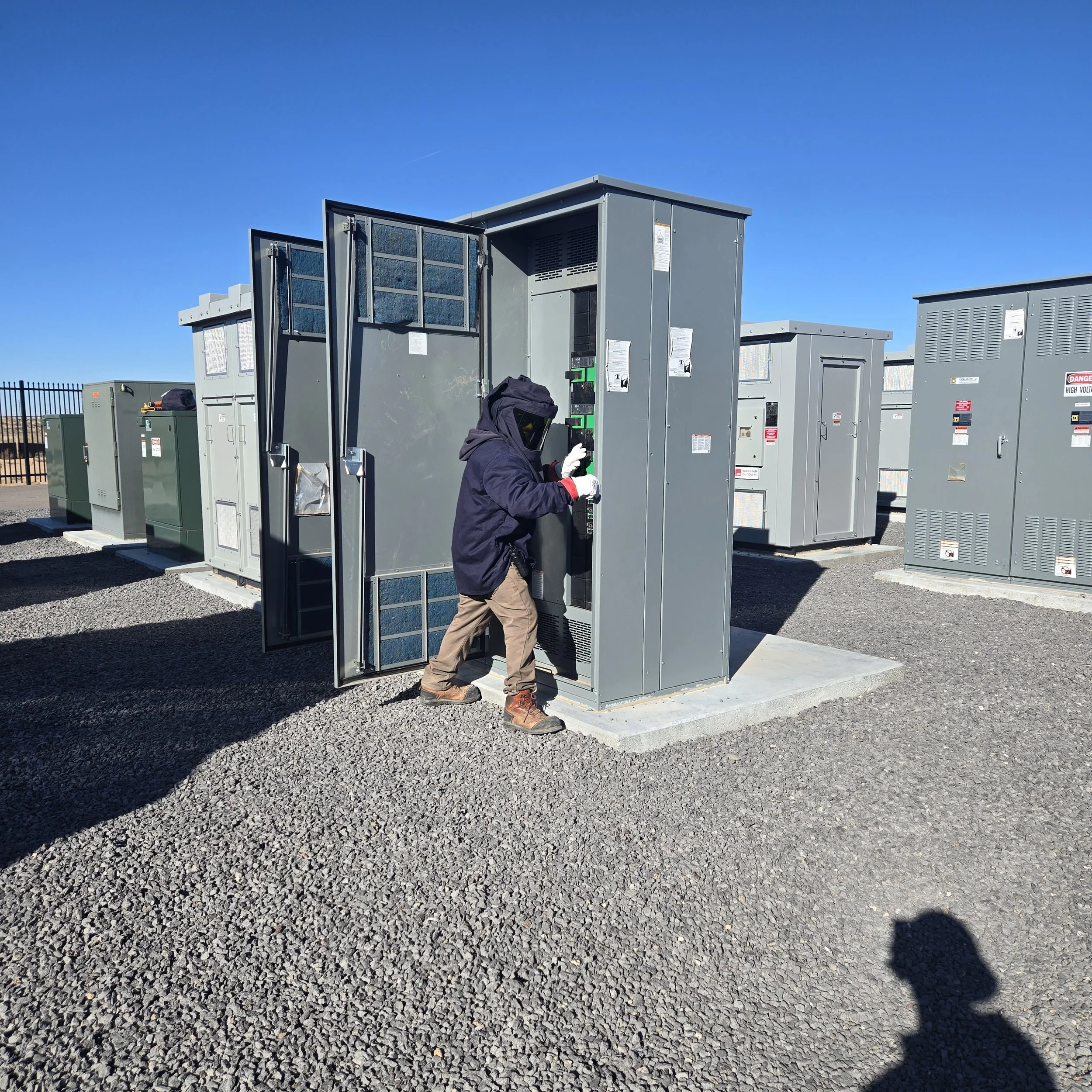 Worker in dark jacket and safety helmet working on electrical equipment at an outdoor industrial site with multiple electrical cabinets and a clear blue sky.