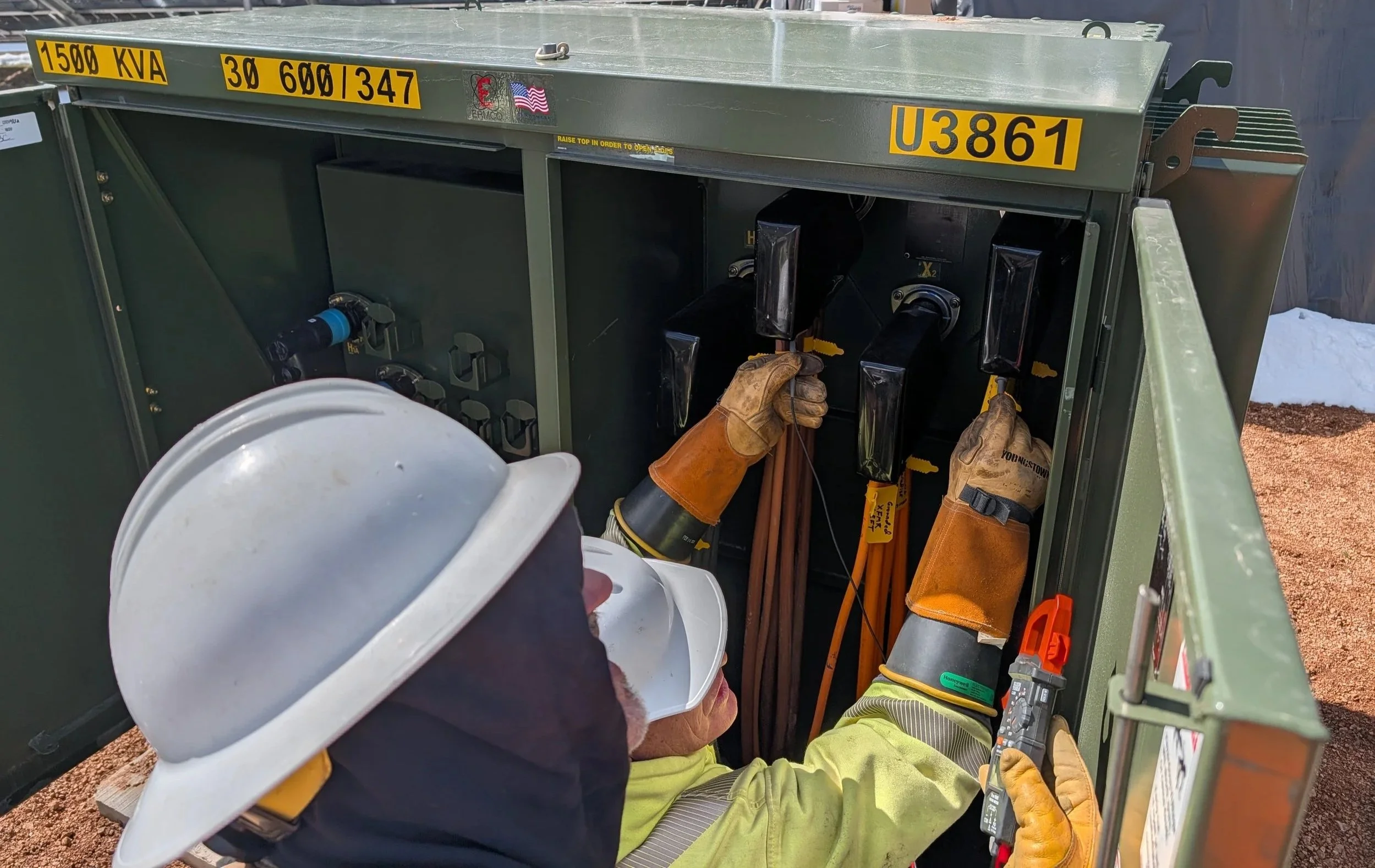 A utility worker wearing a white hard hat and gloves is working inside a green electrical service box with various tools and equipment on a construction site in Crested Butte Colorado.