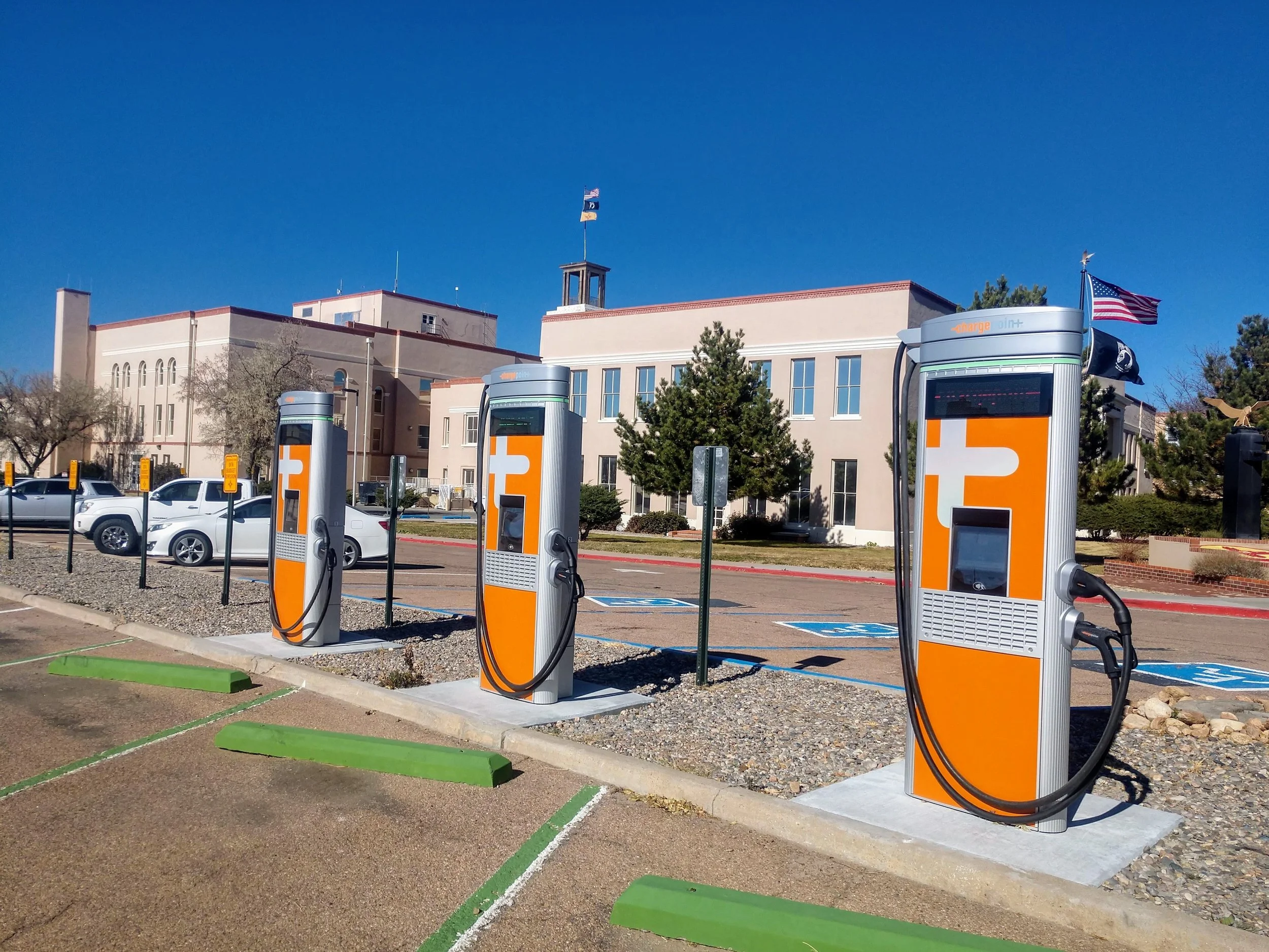 Electric vehicle charging stations in a parking lot with marked spaces and the New Mexico State Capitol Building with American flags in the background.