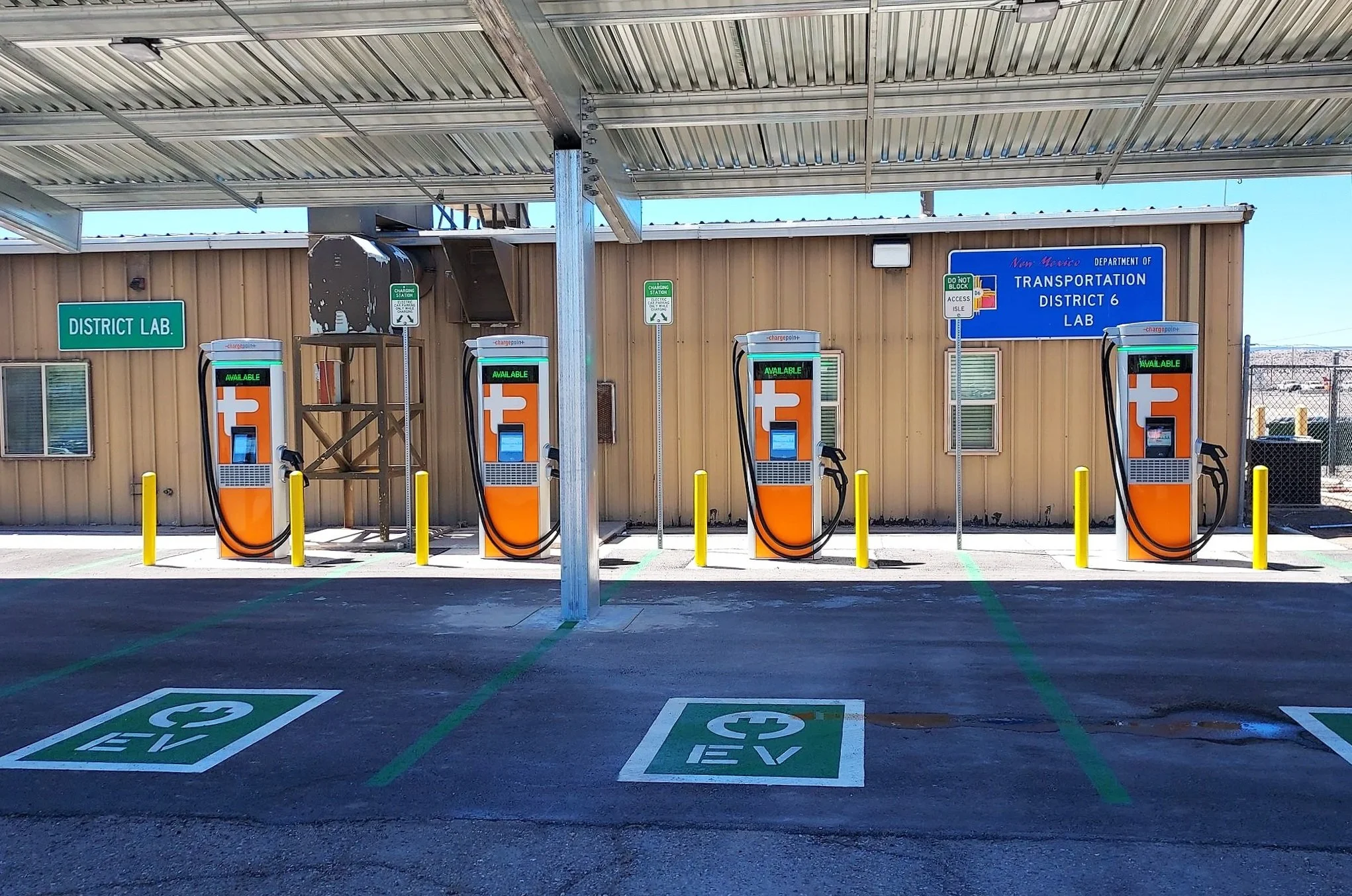 Electric vehicle charging station area with four charging units, parking spaces marked for electric vehicles. A building with signs reading 'District Lab' and New Mexico 'Transportation District 6 Lab' is in the background.