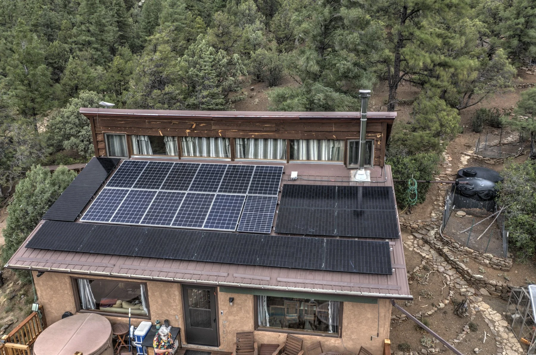 A house with solar panels installed on its roof, an energy storage system, surrounded by trees and woodland, with a patio and outdoor furniture visible in the backyard.