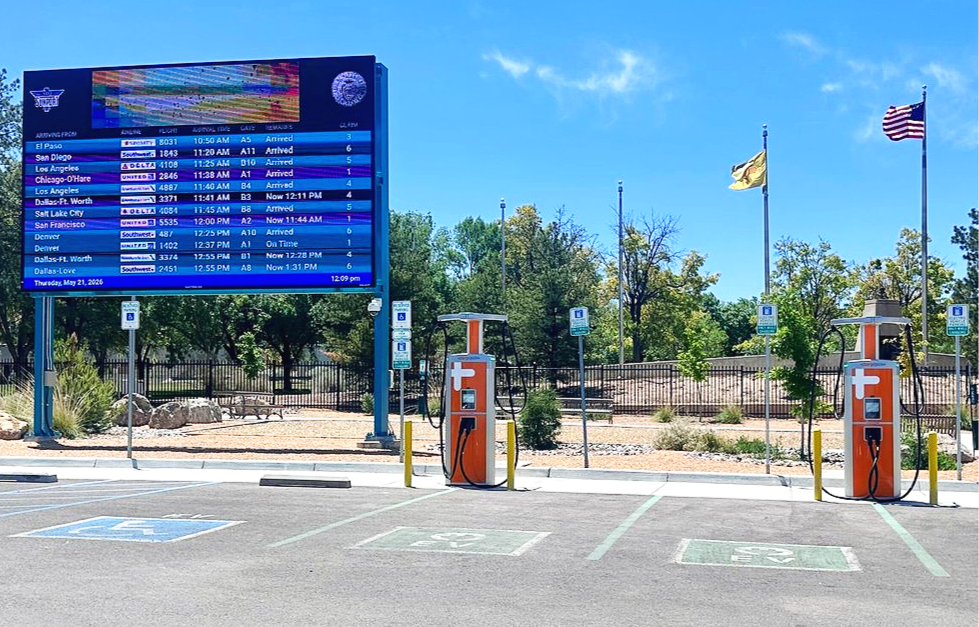 Two electric vehicle charging stations in front of a digital flight arrivals board at the Albuquerque International Sunport airport, with parking spaces marked for handicapped, flags flying in the background on a sunny day.