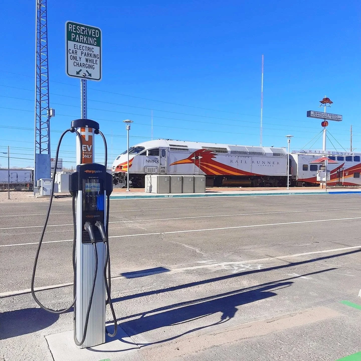 Electric vehicle charging station in parking lot with a rain runner train in the background and blue sky.